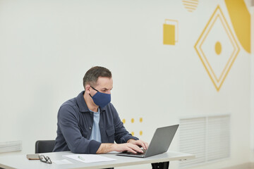 Portrait of mature college professor wearing mask while using laptop in modern school auditorium, copy space