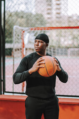Chico negro apuesto posando con balon de baloncesto frente una cancha de baloncesto con reja y edificios de fondo