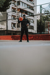 Chico negro apuesto posando con balon de baloncesto frente una cancha de baloncesto con reja y edificios de fondo