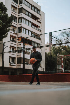 Chico negro apuesto posando con balon de baloncesto frente una cancha de baloncesto con reja y edificios de fondo