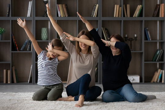 Funny three generations of Caucasian women sit on floor at home make hand gesture sign dab dance together. Overjoyed little girl child with modern young mother and elderly grandmother relax have fun.