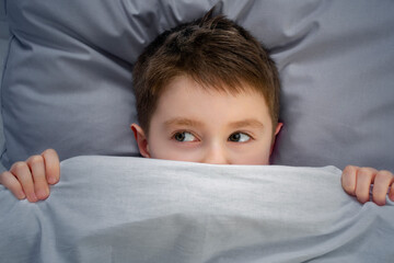 Boy covered his face with a blanket out of fear, the excited child not sleep at night and hides in bed with frightened eyes. Childrens nightmares, fears, imaginations and fantasies in the dark