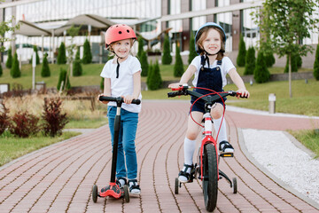 Two little girls having fun on bicycle and scooter. Cheerful sisters in helmets riding outdoors.
