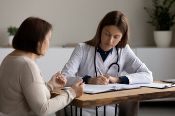 Fototapeta premium Young female doctor or therapist write prescription consult mature patient at meeting in clinic. Caucasian woman GP note complaints in medical journal at consultation with senior client in hospital.