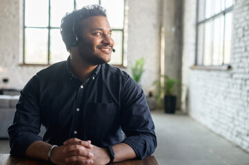 Delighted hindoo man wearing headset device and smart casual shirt looks away dreamily, support representative on the workplace in modern loft office, hindu guy in headphones with mic lost in thoughts
