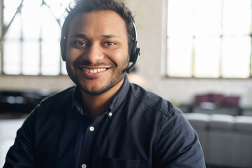 Close-up portrait of cheerful indian guy wearing wireless headset looks at the camera with toothy smile, video connect with multiracial colleague, video chat with hindu guy in headphones, webcam view