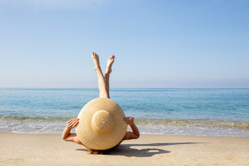 Young woman lying on the beach with legs pointing up to the sky covering her body with broad brim...