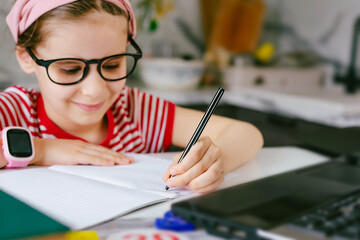 Distance learning online education. Sickness schoolgirl left-handed girl writes with her left hand studying at home with laptop on table doing school homework. Training books and notebooks on table.