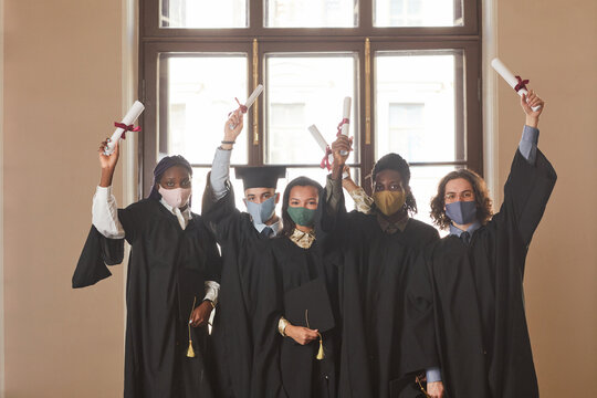 Diverse Group Of Young People Wearing Masks And Black Ceremonial Robes During Indoor Graduation Ceremony In Covid Pandemic
