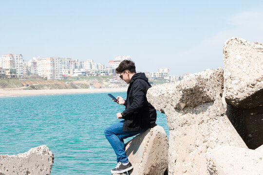 Young man sitting by rock using his mobile phone 