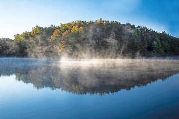 Perfect mirror images of woods reflected in a clear lake. with fog rising. - 431550277