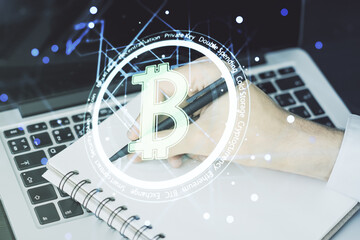 Double exposure of creative Bitcoin symbol hologram with hand writing in notepad on background with laptop. Mining and blockchain concept