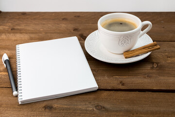 White vintage cup with coffee, espresso with cinnamon sticks on a wooden table. Open notebook and pen. Place for your text and information.