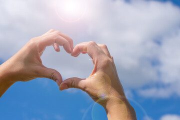 Female young hands in heart shaped gesture on blue cloudy sky background and bright sunlight. Concept of love sicking, relationship, calmness and tranquility. Summer love mood