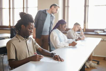 Diverse group of students taking exam in college while sitting in row at desk in auditorium, focus on young African-American man in foreground, copy space
