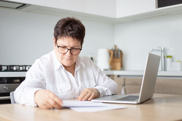 Adult senior 60s woman calculating paper documents at home at pc. Serious middle aged woman at home working on bank payments online with computer technologies.
