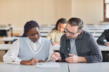 Portrait of mature professor helping African-American woman studying in college auditorium, copy space