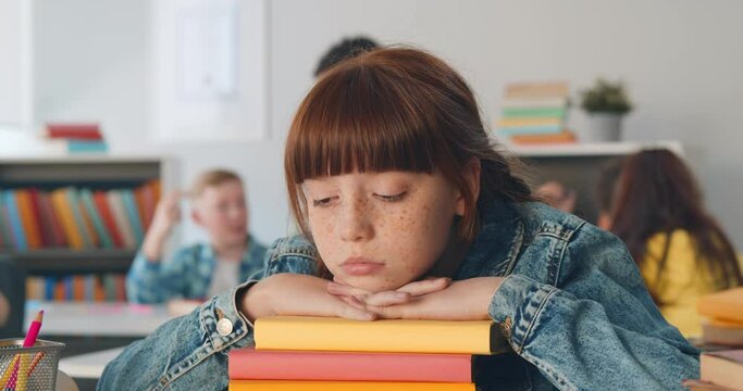 Bored schoolgirl in casualwear keeping her chin on top of stack of books sitting at desk