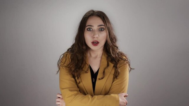 Isolated portrait of woman shocked by some news who is closing her mouth by hand in slowmo. A serious young lady is standing on white background and suddenly getting amazed by unbelievable information