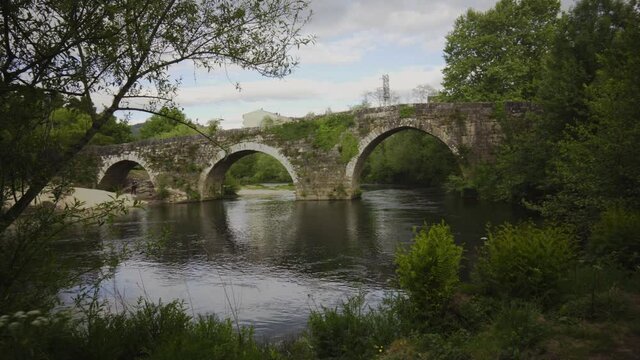 Roman stone bridge that crosses a river. Roman bridge of Ponteareas. stone bridge with green vegetation, cloudy sky and water with reflections, with beach on the shore