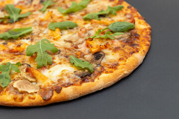 Closeup of a round pizza with cheese, chicken, mushrooms and arugula and spinach on a gray background.