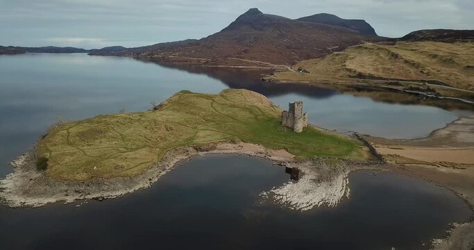 4k aerial footage of ruins of Ardvreck Castle and Loch Assynt with mountains in background. Castle, clouds and sky reflected in calm water of loch. Panning around castle in circular movement.