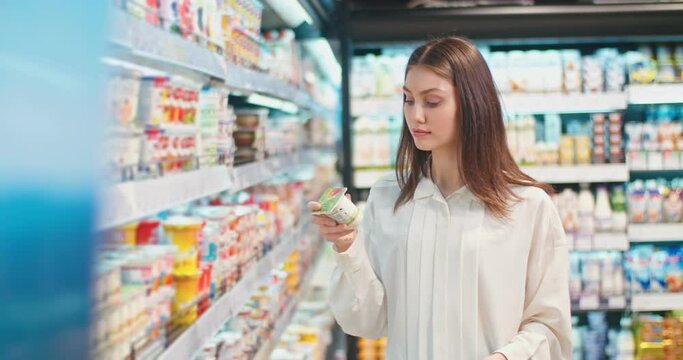 Good-looking young woman choosing yogurt in dairy section at supermarket. Caucasian lady reading composition of goods on package and putting into shopping cart in grocery store. GM products, health.