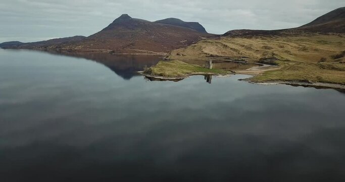 4k aerial footage of ruins of Ardvreck Castle and Loch Assynt with mountains in background. Castle and sky reflected in calm water of loch. Panning backwards away from castle.