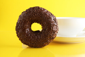 A white cup on a saucer with tea or coffee next to lie cubes of sugar slices and a doughnut in the glaze on a bright yellow background with a place for a copyspace. Still life with sweet food
