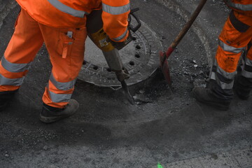 Stra&szlig;enarbeiter mit orangefarbener Hose arbeitet mit Presslufthammer an einem Kanaldeckel