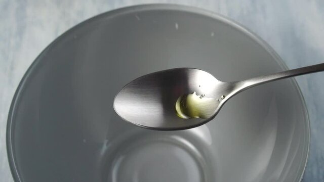 Fresh vegetable oil splashes and overflows a teaspoon and pours into a gray bowl in slow motion. The texture of the wooden kitchen table. Macro shot