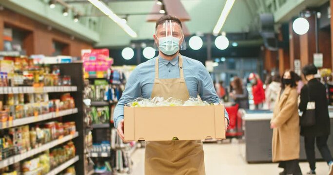 Handsome Caucasian man wearing face visor carrying box with vegetables in supermarket. Male grocery store owner in medical mask walking through products sections in store. Business, virus concept.