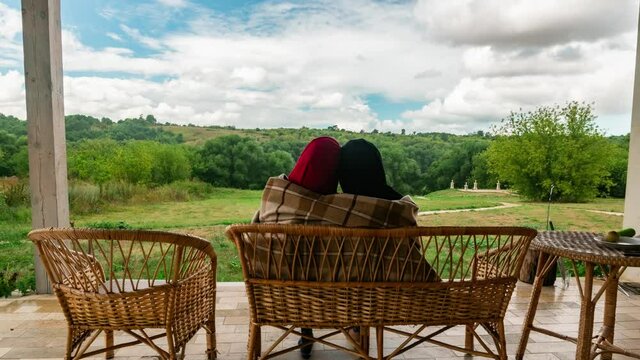 Family couple rests in village house sitting on bench and hugging in timelapse. Man and woman relax on terrace of countryside home covered by blanket. Back view of wife and husband enjoying landscape.