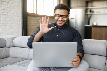 Young cheerful multiethnic guy waving hello to online interlocutor, using laptop computer for virtual video meeting with a family or colleagues, hindu man staying in touch during quarantine