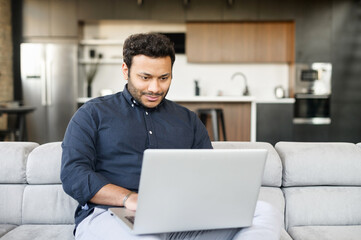 Smiling cheerful mixed-race freelancer guy is using laptop computer sitting on the couch at home, hindu guy enjoys remote work from home in comfy atmosphere. Indian male spends leisure online
