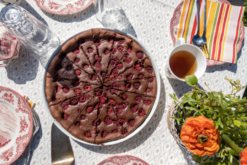 Wonderful summer sweet chocolate cake with cherry. Afternoon teatime with flower and laid table with dish. Self baked chocolate cake on white tablecloth in the outside at daylight