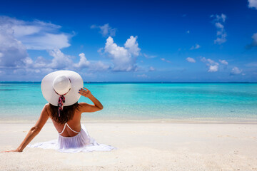 Fototapeta na wymiar A traveler woman with hat sits on a tropical paradise beach and enjoys the view to the turquoise sea
