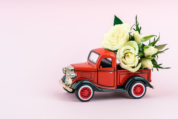 Red toy truck delivering bouquet of white rose flowers on pink background. February 14, Valentine's day, 8 March, International Women's Day. Flower delivery. Copy space.