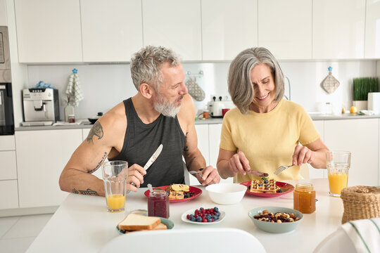 Happy Healthy Fit Mature Older Family Couple Having Breakfast Sitting At Kitchen Table. Smiling Mid Age 50s Husband And Wife Talking While Eating Waffles, Having Fun Enjoying Morning Meal Together.