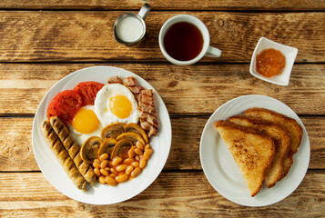 Traditional morning English breakfast - scrambled eggs, beans, tomatoes, mushrooms, sausages and apricot jam with classic English fried toast on a white plate, and a cup of tea with milk - top view