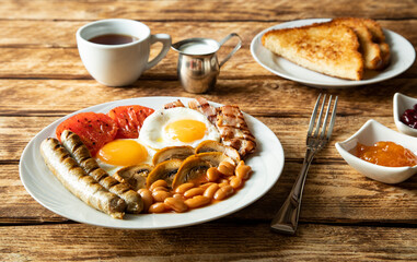 Traditional morning breakfast in England - sausages, tomatoes, eggs, bacon, mushrooms, white beans, toasted toast, a cup of black coffee and jam on a wooden table -  side view and close-up