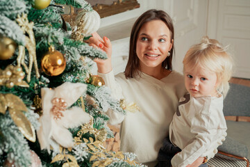 Happy family, brunette mom and baby boy dress up the Christmas tree before Christmas. Beautiful decoration toys on the tree. Modern clothing for the home.