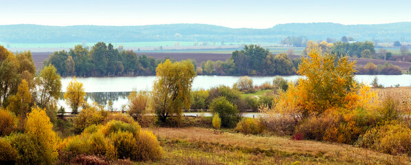 Autumn landscape with colorful trees and bushes on the shore river and forest in the distance, panorama. Golden autumn