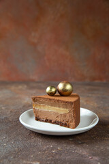 A piece of chocolate cake with a beautiful cut decorated with golden spheres on top. Dessert lies on a white plate on a textured table - close-up, vertical
