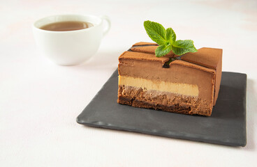 A piece of chocolate modern cake on a square black plate with a leaf of green mint with a beautiful cut and a cup of hot tea - close-up