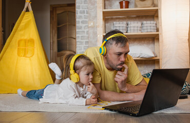 a father and his little daughter are lying on the floor with yellow headphones, looking at a laptop and holding a finger in their mouth.