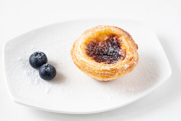 Traditional dessert from Portugal or Japan Pastel de nata on a white plate sprinkled with powdered sugar. The composition is complemented by two fresh blueberries