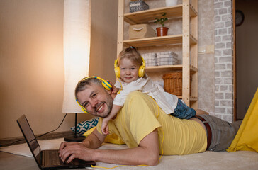 a little girl lies on her father's back and listens to music with yellow headphones while looking at her laptop.