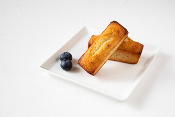 Two French financier cupcakes made of almond flour, butter, almonds, egg white, and powdered sugar on a square white plate with blueberries on the white table