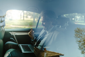 Fototapeta na wymiar Old businessman sitting with laptop at back of car seat and having conference call.
